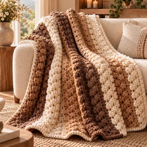 May include: A crochet blanket featuring a striped pattern in shades of cream, light brown, and dark brown. The textile has a textured, bobble-like stitch design throughout, creating a thick, layered appearance as it drapes over a sofa.