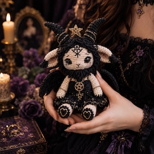 Könnte beinhalten: Eine handgefertigte Häkelpuppe in Schwarz und Creme mit großen dunklen Augen und markanten Hörnern. Die Puppe trägt ein schwarzes Kleid mit goldenen Pentagramm- und Sternanhängern und hat kleine schwarze Flügel an ihrem Rücken befestigt.