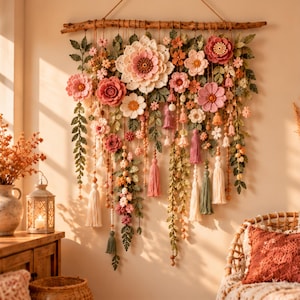 May include: A wall hanging featuring a horizontal wooden branch adorned with numerous crocheted flowers in shades of pink, peach, and white, interspersed with green crocheted leaves, hanging strings of beads, and various fabric tassels.