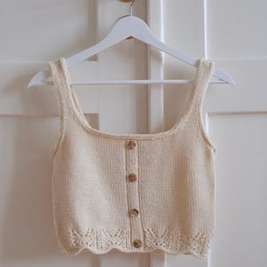 Op de afbeelding: Crèmekleurige gebreide tanktop met een ronde hals en een knoopsluiting aan de voorkant. De mouwloze top heeft een geschulpte zoom en wordt getoond op een witte hanger tegen een witte deur.