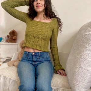 May include: A green, long-sleeved knitted top with a square neckline. The top is worn with blue jeans. The woman is seated on a white sofa, with a decorative cushion and a teddy bear visible in the background.