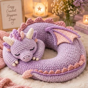 May include: A lavender crochet dragon pillow shaped like a curled-up creature with closed eyes. It features cream-colored horns, a light pink wing, and a tail with small pink triangular spikes along the back. The texture is soft, yarn-based, and plush.