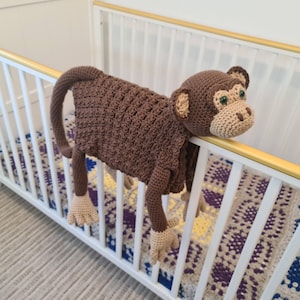 May include: A handmade brown crochet monkey toy with tan accents, hanging on a white crib. The monkey has a long tail, arms, and legs, with green eyes and a tan face. The crib has a gold-colored top rail.