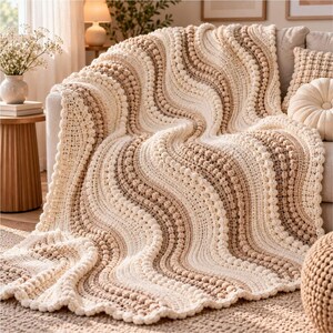 May include: A crochet throw blanket featuring a wavy, rippled pattern in alternating stripes of cream, beige, and light brown. The texture is defined by raised bobble stitch rows that follow the curves, creating a thick, tactile surface.