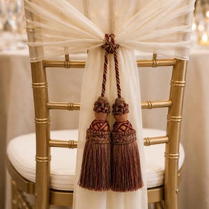 May include: A gold-toned Chiavari chair featuring a white fabric sash draped across the backrest. The sash is secured at the center with a decorative knot and two long, multi-toned brown and copper-colored tassels hanging from braided cords.