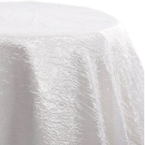 May include: A white, textured tablecloth with a crinkled appearance. The fabric drapes over a round surface, creating soft folds and shadows. The tablecloth is made of a shiny material, reflecting light.