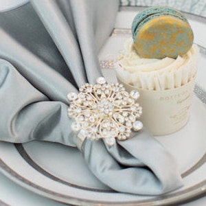 May include: A silver napkin ring with a rhinestone design, holding a light blue napkin. The napkin ring is on a white plate with a silver rim.