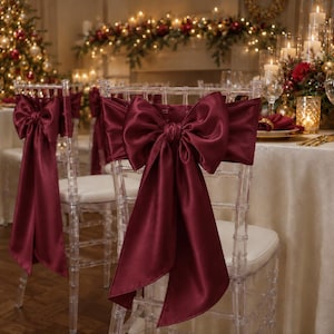 May include: A clear acrylic chair featuring a deep burgundy satin sash tied into a large, prominent bow on the backrest. The fabric has a smooth, reflective finish, and the chair includes a white seat cushion, creating a formal seating arrangement.