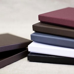 May include: A stack of square, solid-colored boxes in various shades of burgundy, brown, gray, white, and black. The boxes are neatly arranged, with a few boxes lying separately in the foreground. The boxes appear to be made of a sturdy material.