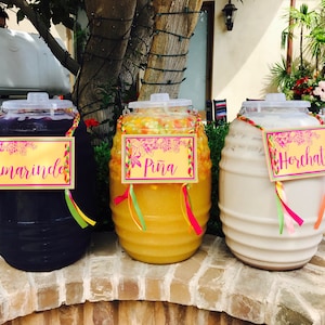May include: Three large glass jars filled with different colored beverages. The jars are labeled "Tamarindo", "Piña", and "Horchata". The jars are decorated with colorful ribbons and flowers.