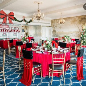 May include: A festive dining room decorated for Christmas with red tablecloths, gold chairs, and black chair covers. The room features a Christmas tree, decorative garlands, and a backdrop with a bow and the text "Gimmit's Desserts."