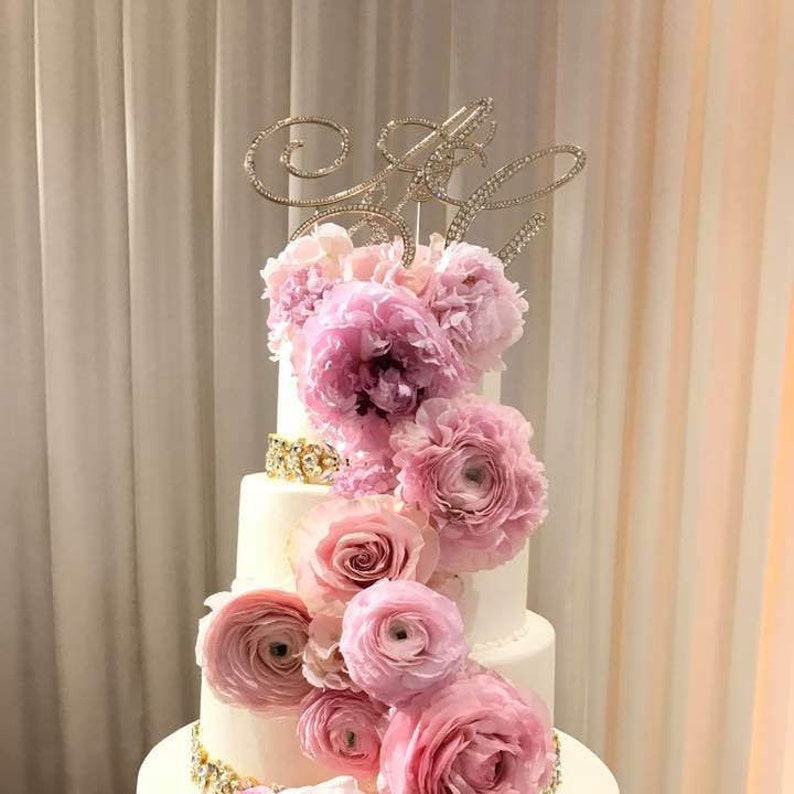 May include: A white wedding cake with four tiers decorated with pink flowers and a gold and crystal topper that says "&".