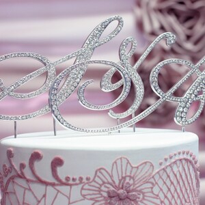 May include: A silver cake topper with the word "Mrs" in cursive, decorated with rhinestones. The topper is on a white cake with pink lace detailing.