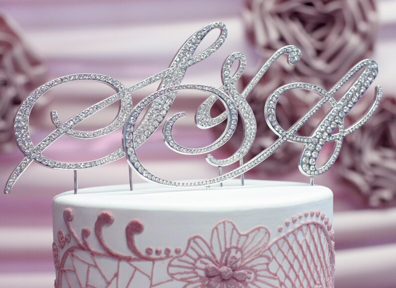 May include: A silver cake topper with the word "Mrs" in cursive, decorated with rhinestones. The topper is on a white cake with pink lace detailing.