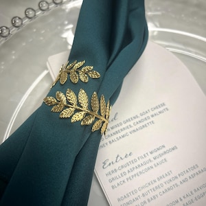 May include: A teal napkin is held in place by a gold-colored leaf-shaped napkin ring. A menu with the word "Entrée" is visible, listing dishes like herb-crusted filet mignon and roasted chicken breast. The napkin is placed on a clear plate.