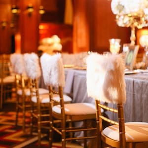 May include: Gold chiavari chairs with white faux fur chair covers lined up in a row. The chairs are set up in a room with a wooden floor and a patterned carpet.