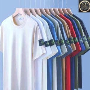 Peut inclure: Une rangée de t-shirts à manches courtes de différentes couleurs, dont crème, blanc, bleu et rouge, suspendus à des cintres en bois. Chaque chemise a un écusson sur la manche gauche. Les chemises sont présentées sur un fond bleu clair.