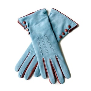 May include: A pair of light blue gloves featuring a soft texture, accented with dark brown leather piping along the fingers and cuffs. Each glove is decorated with three small, round, dark brown buttons on the outer wrist and three vertical stitched lines.