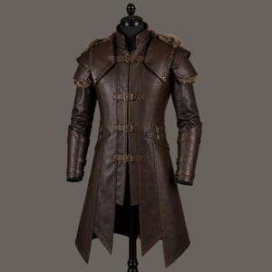 May include: A brown leather coat with a high collar, shoulder pads, and fur trim. The coat features multiple buckles and a long, flowing design. The sleeves have armored detailing, and the coat is displayed on a black mannequin.