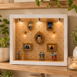 May include: A white framed shadow box featuring miniature framed art and figures. The art includes a cat portrait and landscape scenes. The box is lit with small spotlights and decorative light fixtures. The background is a patterned wallpaper.
