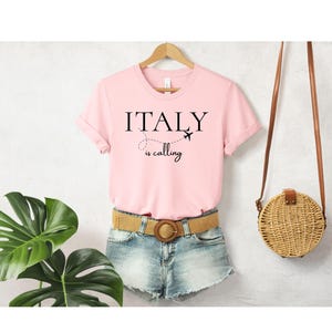 Könnte beinhalten: Ein hellrosa kurzärmeliges T-Shirt mit dem schwarzen Schriftzug ITALY is calling, einem kleinen Flugzeugsymbol und einer gepunkteten Flugroute. Das T-Shirt hängt an einem Holzkleiderbügel und wird mit hellblauen Jeansshorts kombiniert.