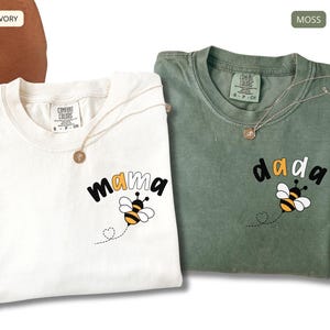 May include: An ivory t-shirt with the word mama and a bee graphic, and a moss green t-shirt with the word dada and a bee graphic. Both shirts feature a Comfort Colors label at the neck and a small bee illustration with a dotted flight path.