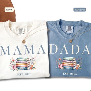 May include: Two crewneck t-shirts in ivory and blue jean colors. The ivory shirt features the text MAMA and the blue jean shirt features DADA, both with a stack of colorful books, floral illustrations, and the text EST. 2026 printed below.