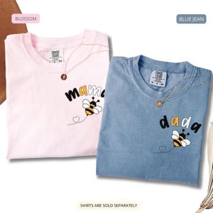 May include: Two short-sleeve crew neck t-shirts in blossom pink and blue jean colors. Each shirt features a graphic of a bee with a dotted flight path forming a heart, and the text mama on the pink shirt and dada on the blue shirt.