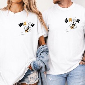 May include: Two white short-sleeve t-shirts featuring a small graphic on the left chest. One shirt displays the text "mama" with a bee illustration, and the other displays the text "dada" with a bee illustration. Both include a dotted heart trail.