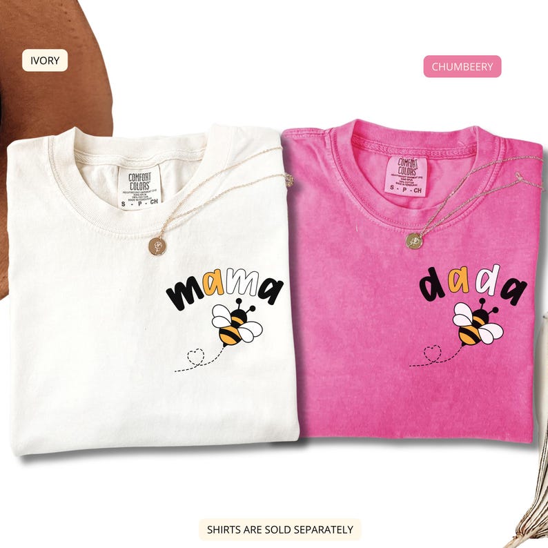 May include: Two short-sleeve crew neck t-shirts, one ivory and one pink, each featuring a graphic of a bee and the text 'mama' or 'dada' on the chest. The shirts have a Comfort Colors brand label inside the collar and a dotted heart trail.