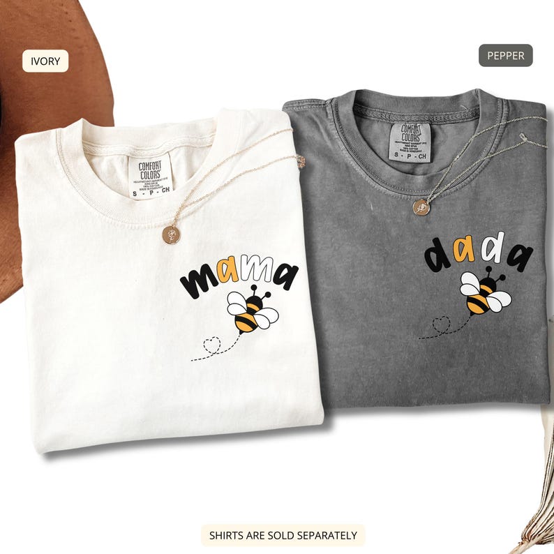 May include: Two short-sleeve crew neck t-shirts, one ivory and one pepper gray, each featuring a small graphic on the left chest. The ivory shirt displays the word mama in black with a bee illustration, while the gray shirt displays the word dada in black with a bee illustration.