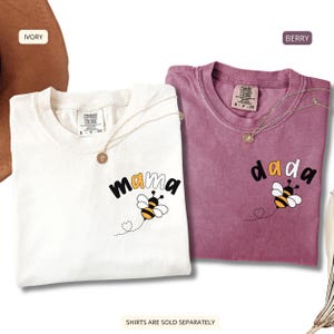 May include: An ivory t-shirt with the word mama and a bee graphic, and a berry t-shirt with the word dada and a bee graphic. Both shirts feature a Comfort Colors label at the neck and a small gold-toned necklace resting on the collar area.