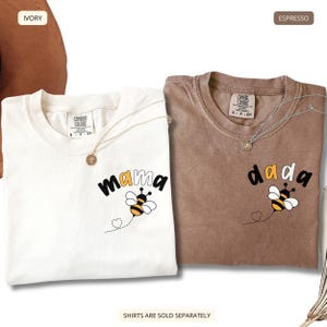 May include: Two t-shirts, one ivory and one espresso, each featuring a graphic of a bee and the text 'mama' or 'dada'. The shirts have a crew neckline and a Comfort Colors brand label at the inner collar. A thin silver necklace rests on each shirt.