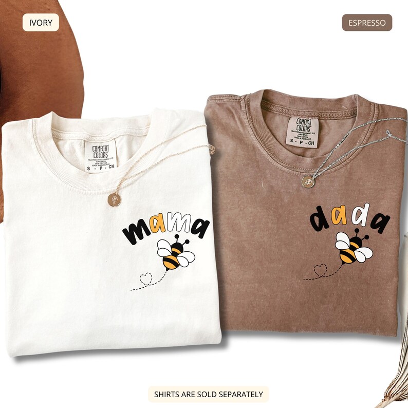 May include: Two t-shirts, one ivory and one espresso, each featuring a graphic of a bee and the text 'mama' or 'dada'. The shirts have a crew neckline and a Comfort Colors brand label at the inner collar. A thin silver necklace rests on each shirt.