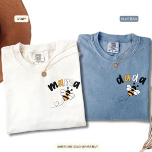 May include: Two short-sleeved t-shirts, one ivory and one blue jean, each featuring a graphic of a bee and the text 'mama' or 'dada' on the chest. The shirts have a Comfort Colors label at the inner neckline and a small heart-shaped flight path.