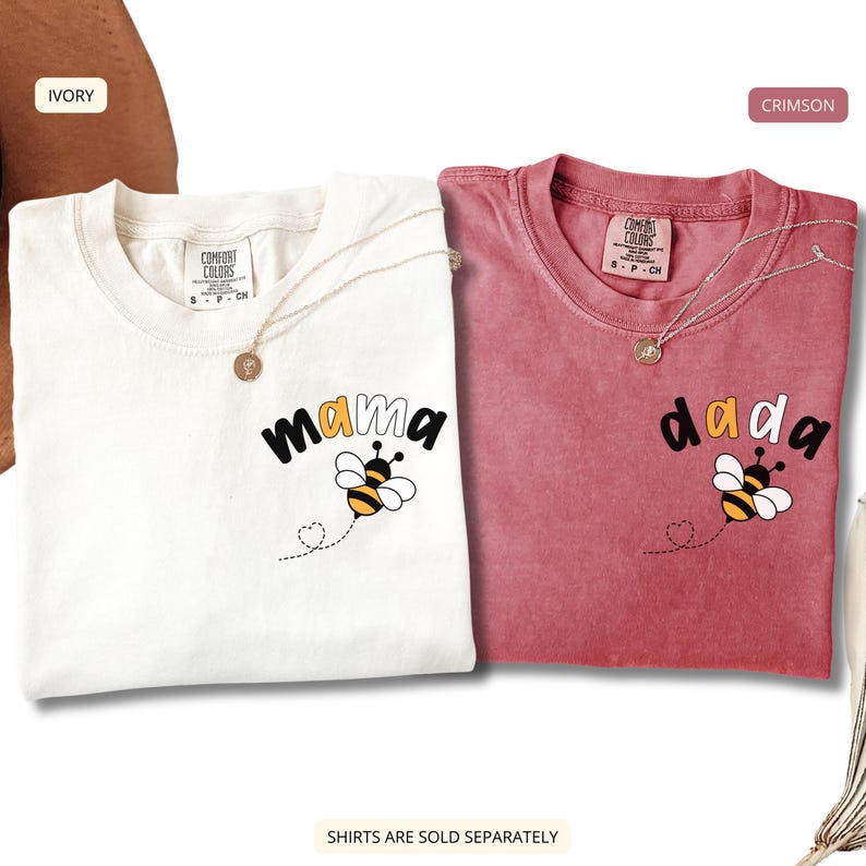 May include: Two short-sleeve crew neck t-shirts, one ivory and one crimson, each featuring a graphic of a bumblebee with a dotted flight path ending in a heart. The ivory shirt has the text 'mama' and the crimson shirt has the text 'dada'.