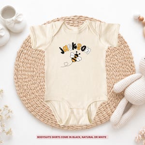 May include: A natural-colored short-sleeve bodysuit featuring the name Jackson in black and orange lettering above a graphic of a bumblebee with a dotted flight path. The text at the bottom states: Bodysuits shirts come in black, natural or white.