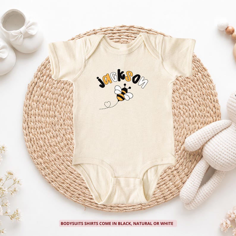 May include: A natural-colored short-sleeve bodysuit featuring the name Jackson in black and orange lettering above a graphic of a bumblebee with a dotted flight path. The text at the bottom states: Bodysuits shirts come in black, natural or white.