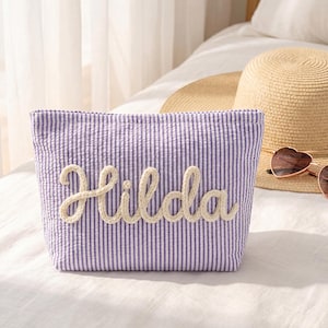 Può includere: Un beauty case a righe lavanda e bianche con il nome "Hilda" ricamato in una scritta color crema. Un cappello di paglia e occhiali da sole a forma di cuore sono sullo sfondo. La borsa è realizzata in tessuto strutturato.
