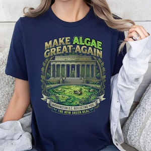 Könnte beinhalten: Ein marineblaues T-Shirt mit einer Grafik des Lincoln Memorials und einem mit grünen Algen gefüllten Reflexionsbecken. Der Text lautet: MAKE ALGAE GREAT AGAIN, WASHINGTON D.C. REFLECTING POOL, THE NEW GREEN DEAL. Das Design zeigt Enten und Laub.