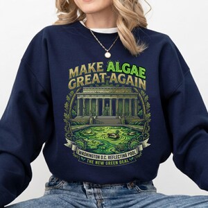 May include: A navy blue long-sleeve sweatshirt featuring a graphic print of the Lincoln Memorial and a green algae-filled reflecting pool. The text reads "MAKE ALGAE GREAT AGAIN" and "WASHINGTON D.C. REFLECTING POOL THE NEW GREEN DEAL" in yellow and green.