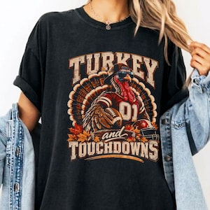May include: A black short-sleeve t-shirt featuring a graphic of a turkey wearing sunglasses and a football jersey with the number 01, holding a football. The text reads TURKEY and TOUCHDOWNS, surrounded by autumn leaves and a football helmet.
