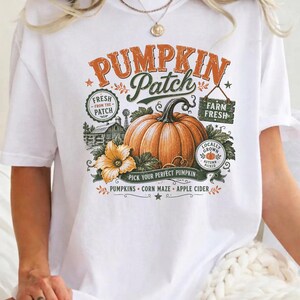 May include: A white t-shirt featuring a graphic print with the text PUMPKIN Patch, Fresh From The Patch, Farm Fresh, Pick Your Perfect Pumpkin, Pumpkins, Corn Maze, and Apple Cider. The design includes a large orange pumpkin and a yellow flower.