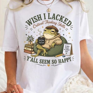 May include: A white t-shirt featuring a graphic of a frog wearing a brown hat and hoodie, sitting in a chair with a mug, books, and a notepad. The text reads: WISH I LACKED Critical Thinking Skills Y'ALL SEEM SO HAPPY. The notepad says: TOO MUCH THOUGHT... NOT ENOUGH PEACE.