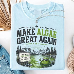May include: A light blue t-shirt featuring a graphic with the text "MAKE ALGAE GREAT AGAIN" in black and green. The design includes a mountain landscape, a lake, a sign reading "CLEAN WATER. GREEN FUTURE. ALGAE IS THE ANSWER.", and a cartoon algae character.