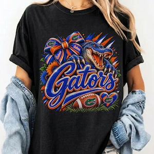 May include: A black short-sleeve t-shirt featuring a graphic print of an alligator, a blue and orange bow, a football, and the word Gators in blue script. The design includes orange and blue accents, sunflowers, and a heart-shaped emblem.