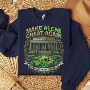 May include: A navy blue crewneck sweatshirt featuring a graphic print of the Lincoln Memorial and a reflecting pool filled with green algae, frogs, and ducks. The text reads "MAKE ALGAE GREAT AGAIN" and "WASHINGTON D.C. REFLECTING POOL THE NEW GREEN DEAL."