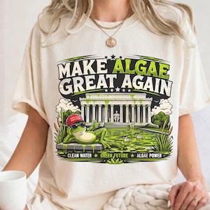 May include: A cream-coloured t-shirt featuring a graphic of a frog wearing sunglasses and a red cap, lounging by a pond in front of a monument. The text reads "MAKE ALGAE GREAT AGAIN" at the top and "CLEAN WATER GREEN FUTURE ALGAE POWER" at the bottom.