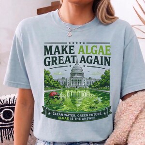 May include: A light blue t-shirt featuring a graphic of the U.S. Capitol building reflected in a green pond. A cartoon algae character wearing a red hat and sunglasses gives a thumbs up. Text reads: MAKE ALGAE GREAT AGAIN, CLEAN WATER. GREEN FUTURE. ALGAE IS THE ANSWER.