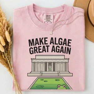 May include: A light pink crew neck t-shirt featuring the black text "MAKE ALGAE GREAT AGAIN" above a graphic of the Lincoln Memorial and a green pond with a frog. The shirt has a Comfort Colors brand label on the inside of the collar.